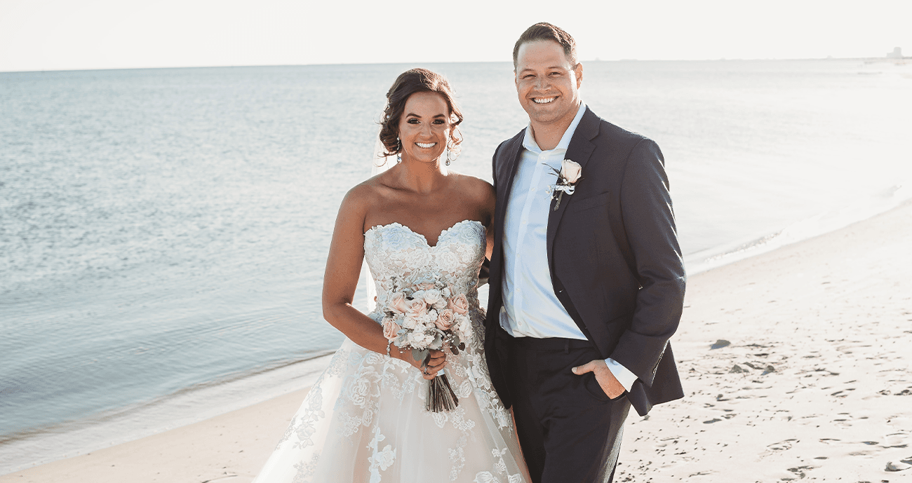 Brian and Kaci at their wedding on a beach together. Kaci is wearing a strapless dress and Brian is wearing a tux.