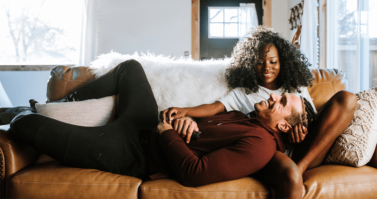 A couple lounges on a sofa, smiling and relaxed—visually capturing the difference between dating vs relationship through their comfort and emotional closeness.