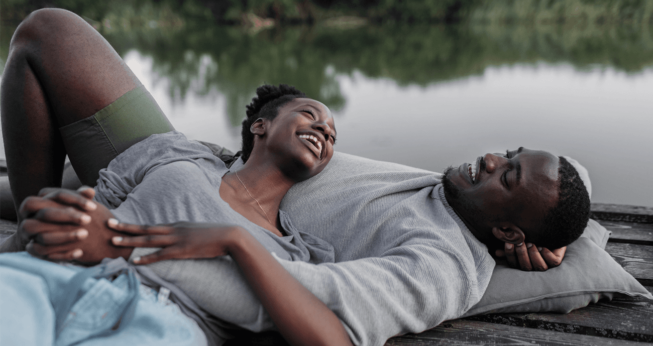 A couple lies on a wooden dock by a calm lake, laughing and holding hands, showcasing relaxed and jhappy relationship dynamics.