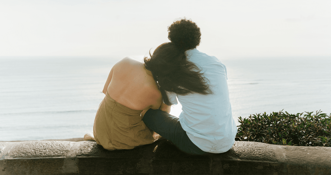 Couple sitting together, looking out at the sea. The woman has her head on the man's shoulder.